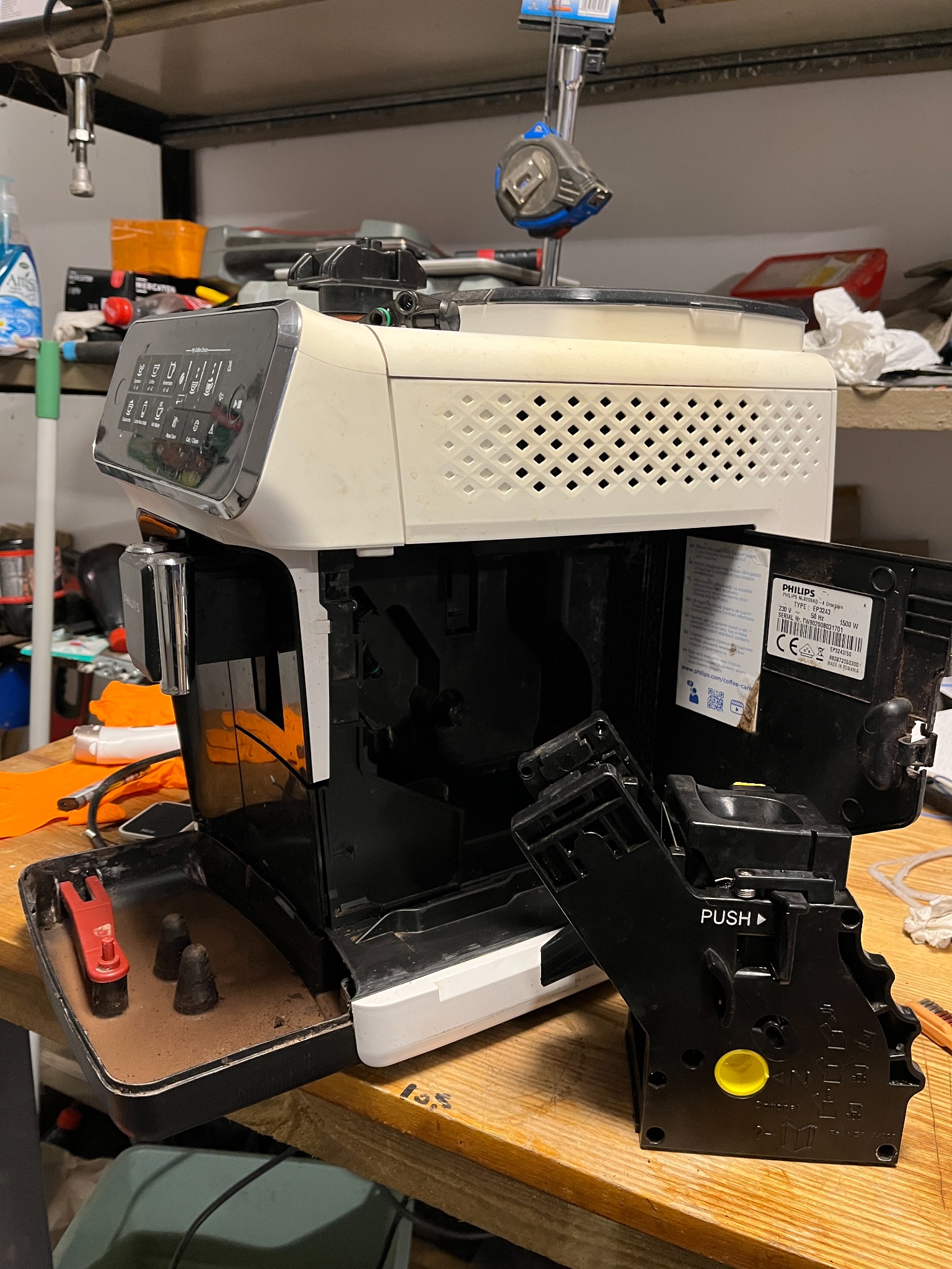 Rozłożony na części ekspres do kawy Philips EP3243 na warsztatowym stole, widoczny otwarty pojemnik na fusy z resztkami kawy oraz wyjęty blok zaparzający z napisem PUSH, w tle półki warsztatowe...