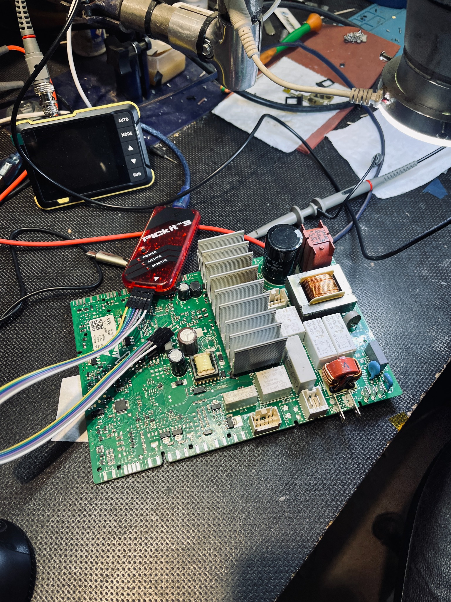 Zielona płytka drukowana z elementami elektronicznymi, podłączona do programatora Pickit 3 i oscyloskopu, leżąca na czarnym, teksturowanym blacie roboczym w warsztacie elektronika.
