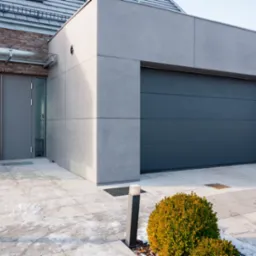 Nowoczesna, segmentowa brama garażowa w kolorze antracytowym, zlicowana ze ścianą budynku o szarej elewacji. Widoczny fragment posesji z kostki brukowej i krzewem ozdobnym. Minimalistyczny styl.