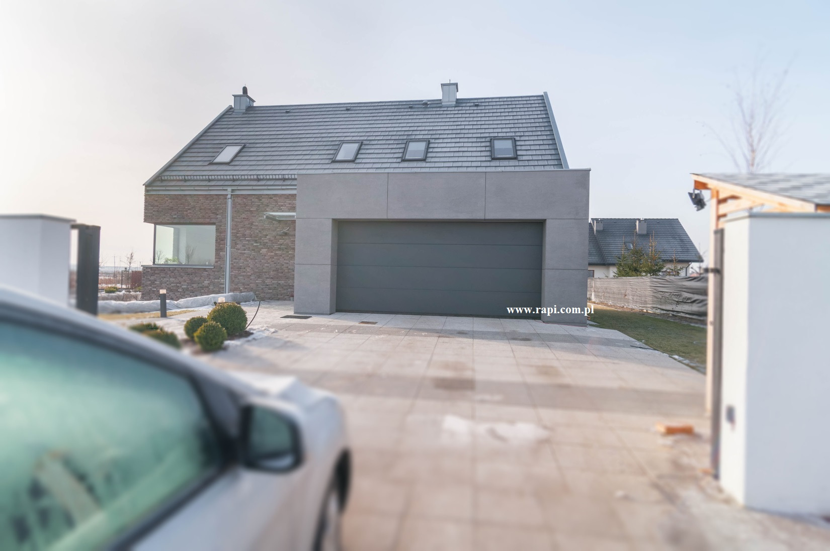 Nowoczesna, szara brama garażowa segmentowa w bryle domu z ceglaną elewacją i szarym dachem, widok z podjazdu z częściowo widocznym samochodem.