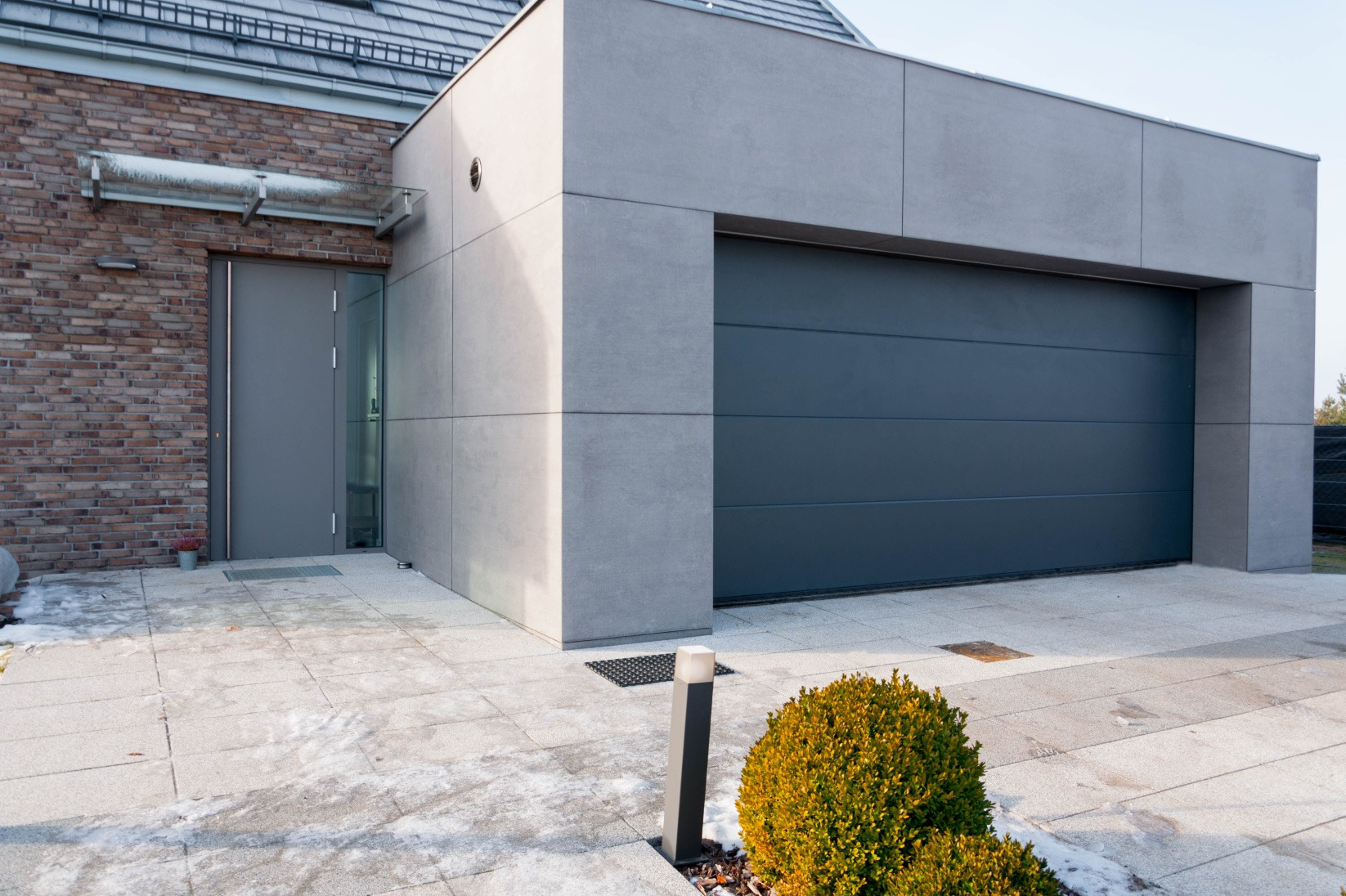 Nowoczesna, segmentowa brama garażowa w kolorze antracytowym, zlicowana ze ścianą budynku o szarej elewacji. Widoczny fragment posesji z kostki brukowej i krzewem ozdobnym. Minimalistyczny styl.