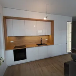 Nowoczesna kuchnia z białymi, lakierowanymi frontami szafek i drewnianym blatem, oświetlona listwą LED podszafkową, czarny zlew i bateria, widok na salon z szarą ścianą dekoracyjną i drabiną.