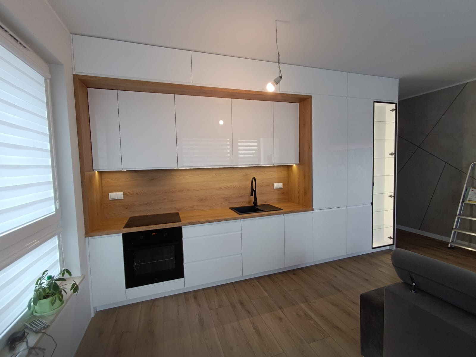 Nowoczesna kuchnia z białymi, lakierowanymi frontami szafek i drewnianym blatem, oświetlona listwą LED podszafkową, czarny zlew i bateria, widok na salon z szarą ścianą dekoracyjną i drabiną.