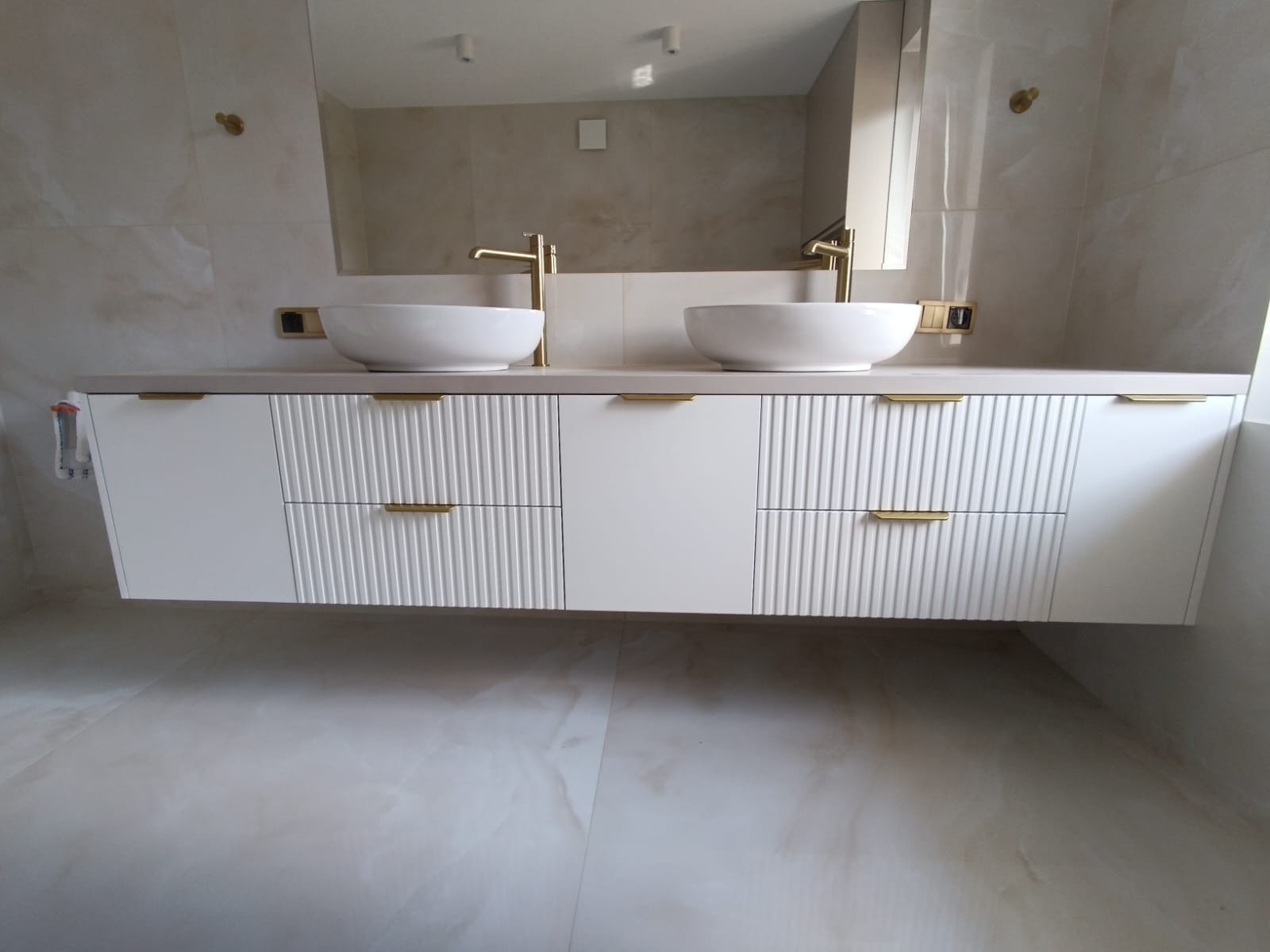 Nowoczesna szafka łazienkowa z dwiema umywalkami nablatowymi i złotymi bateriami, wykonana na wymiar w Kielcach. Fronty szafek posiadają dekoracyjne, pionowe frezowania.