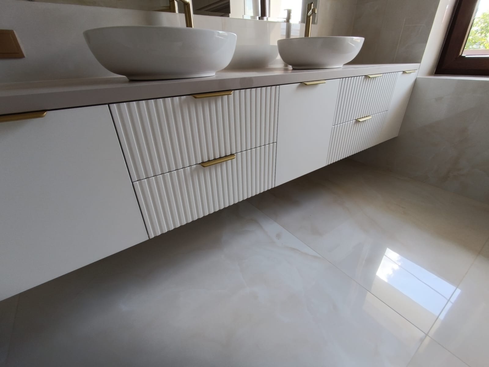 Nowoczesna łazienka z białymi szafkami pod umywalki nablatowe, z frezowanymi frontami i złotymi uchwytami, na tle płytek imitujących marmur.