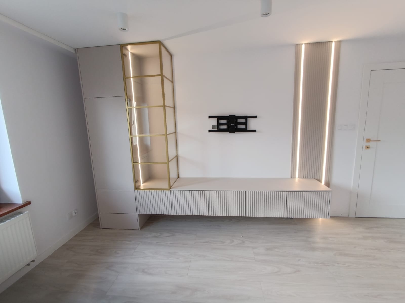 Nowoczesna zabudowa salonu: szafa z przeszkloną witryną z oświetleniem LED, szafka RTV z ryflowanymi frontami, dekoracyjne panele ścienne z podświetleniem oraz uchwyt do telewizora na białej ścianie.