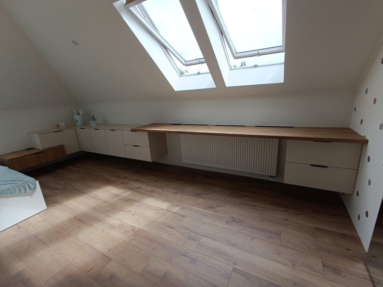 Pokój na poddaszu z zabudową meblową pod skosami, obejmującą szafki z jasnymi frontami i długi, drewniany blat roboczy nad grzejnikiem. Dwa okna dachowe wpuszczają światło.
