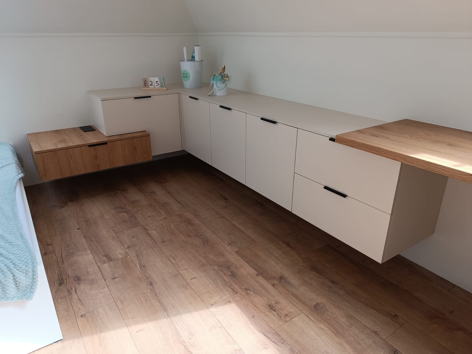 Białe szafki z czarnymi uchwytami i drewnianym blatem wkomponowane w skos dachu, uzupełnione drewnianą szafką nocną, na podłodze panele imitujące drewno.