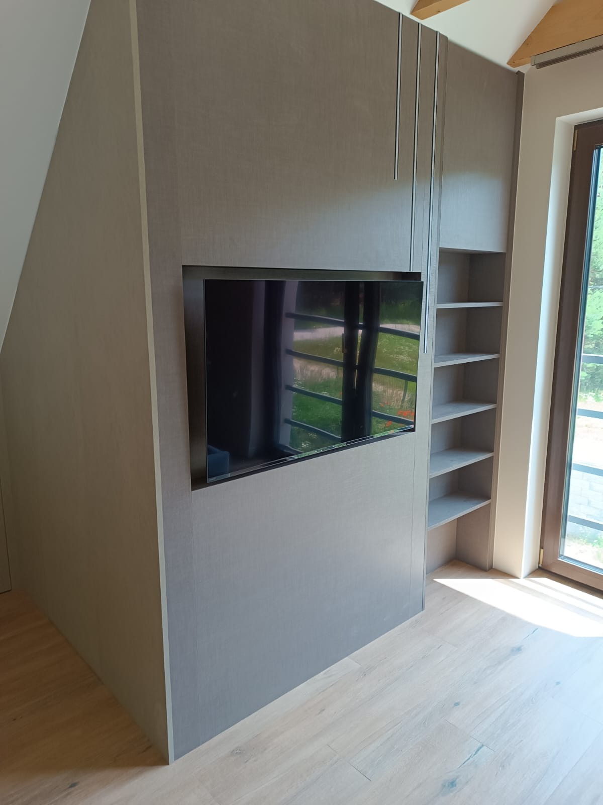 Szara szafa na wymiar z wnęką na telewizor i półkami, dopasowana do skosu dachu, z długimi, pionowymi uchwytami.