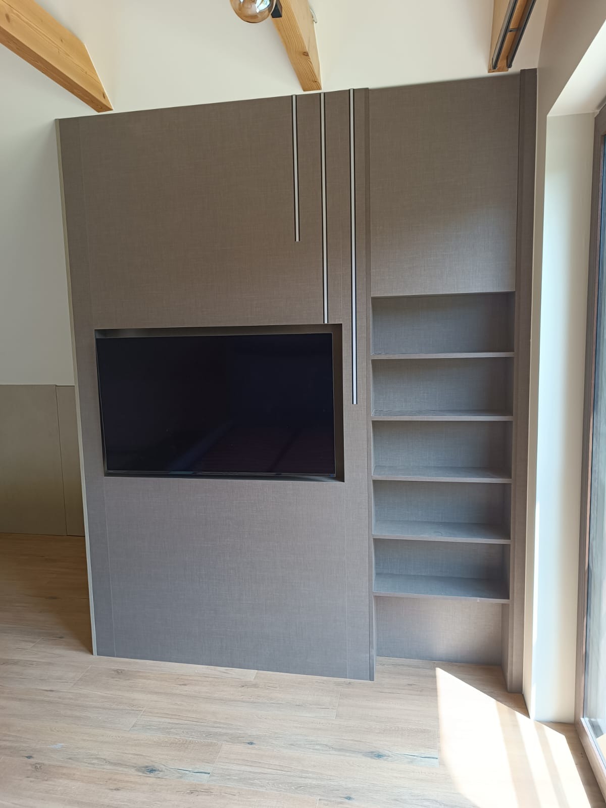 Szara zabudowa meblowa z wnęką na telewizor i regałem na książki, widoczne drewniane belki sufitowe i parkiet na podłodze.