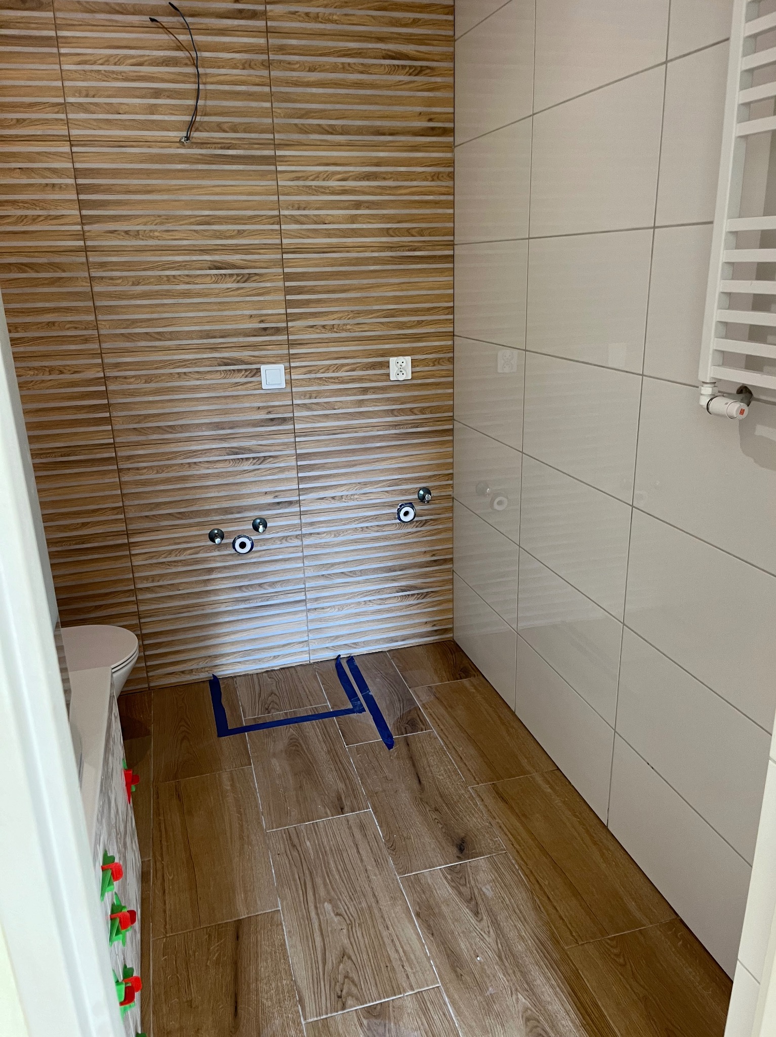 Wykończona łazienka z drewnopodobnymi płytkami na ścianach i podłodze. Widoczne przyłącza wodne, białe płytki na ścianie i grzejnik. Użyto poziomowania płytek.