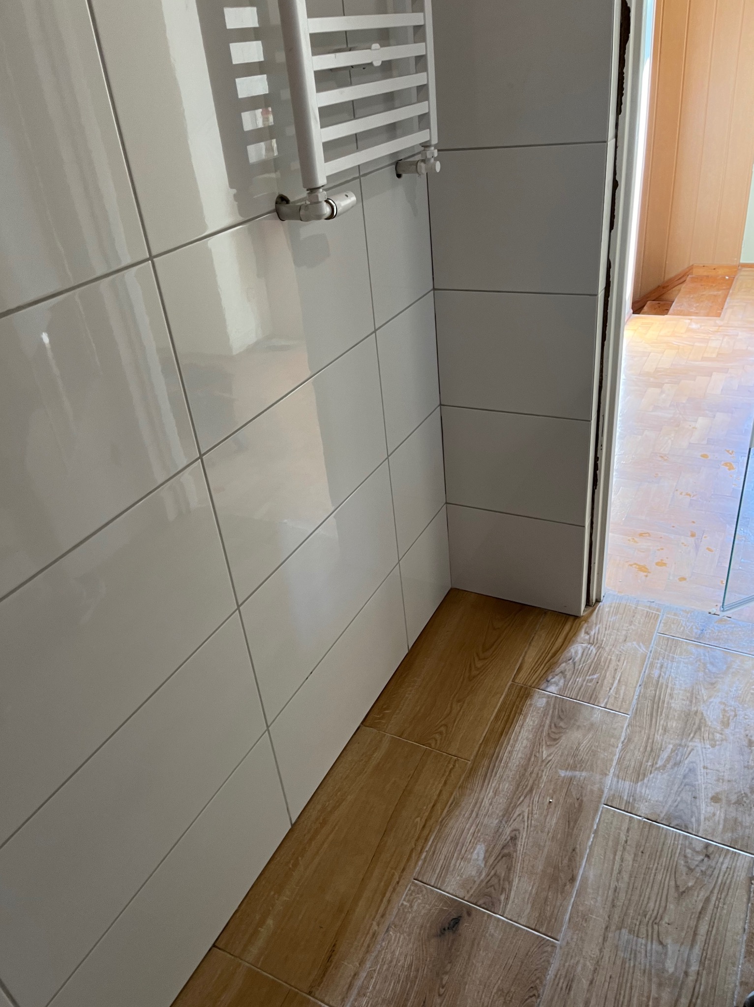 Białe, błyszczące płytki ścienne w łazience, z widocznym grzejnikiem drabinkowym i podłogą imitującą drewno. Widok na sąsiednie pomieszczenie przez otwarte drzwi.