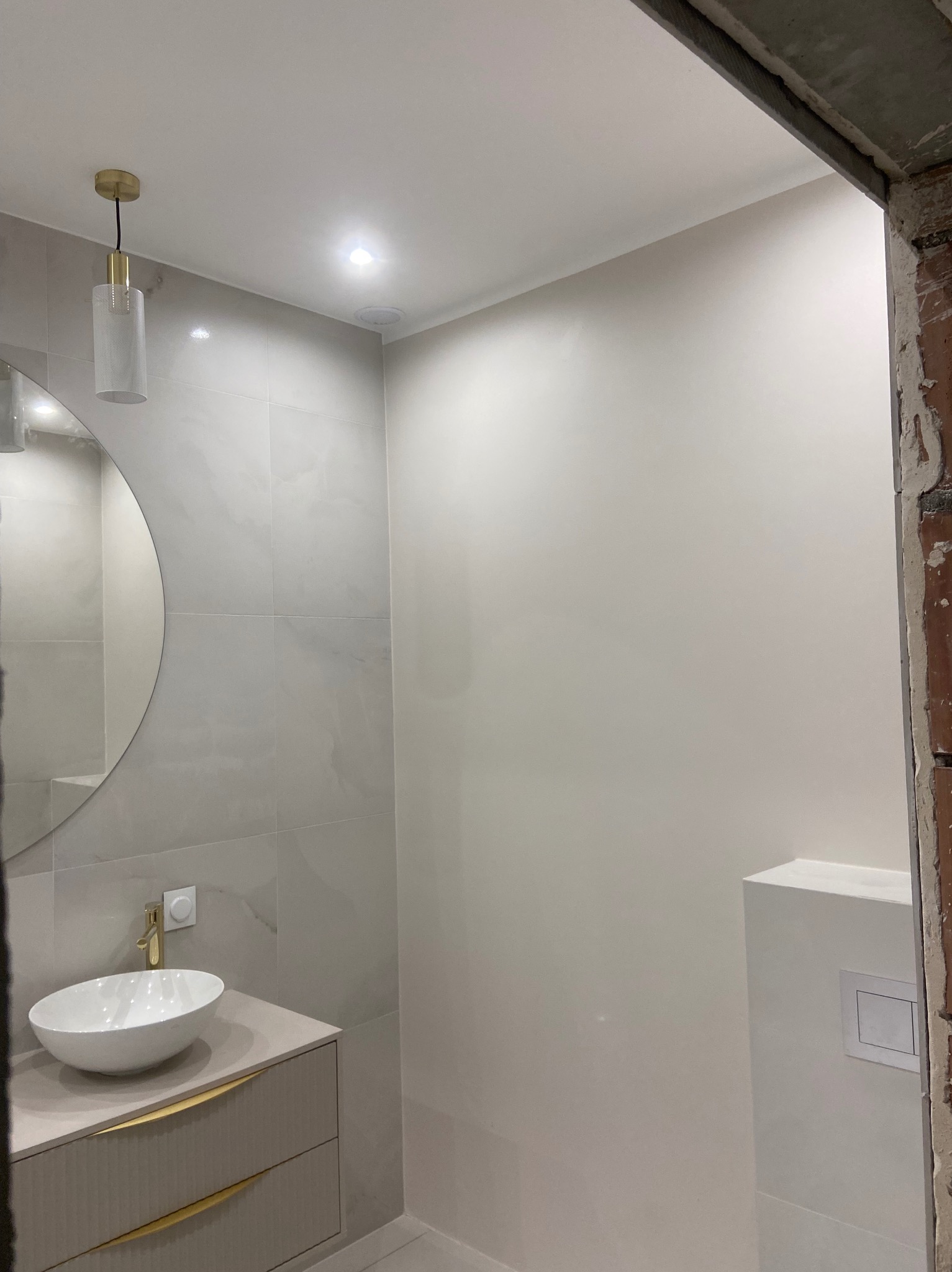 Wykończona łazienka w odcieniach szarości z okrągłym lustrem, złotą armaturą i wiszącą lampą. Widoczny fragment surowego muru przy wejściu.