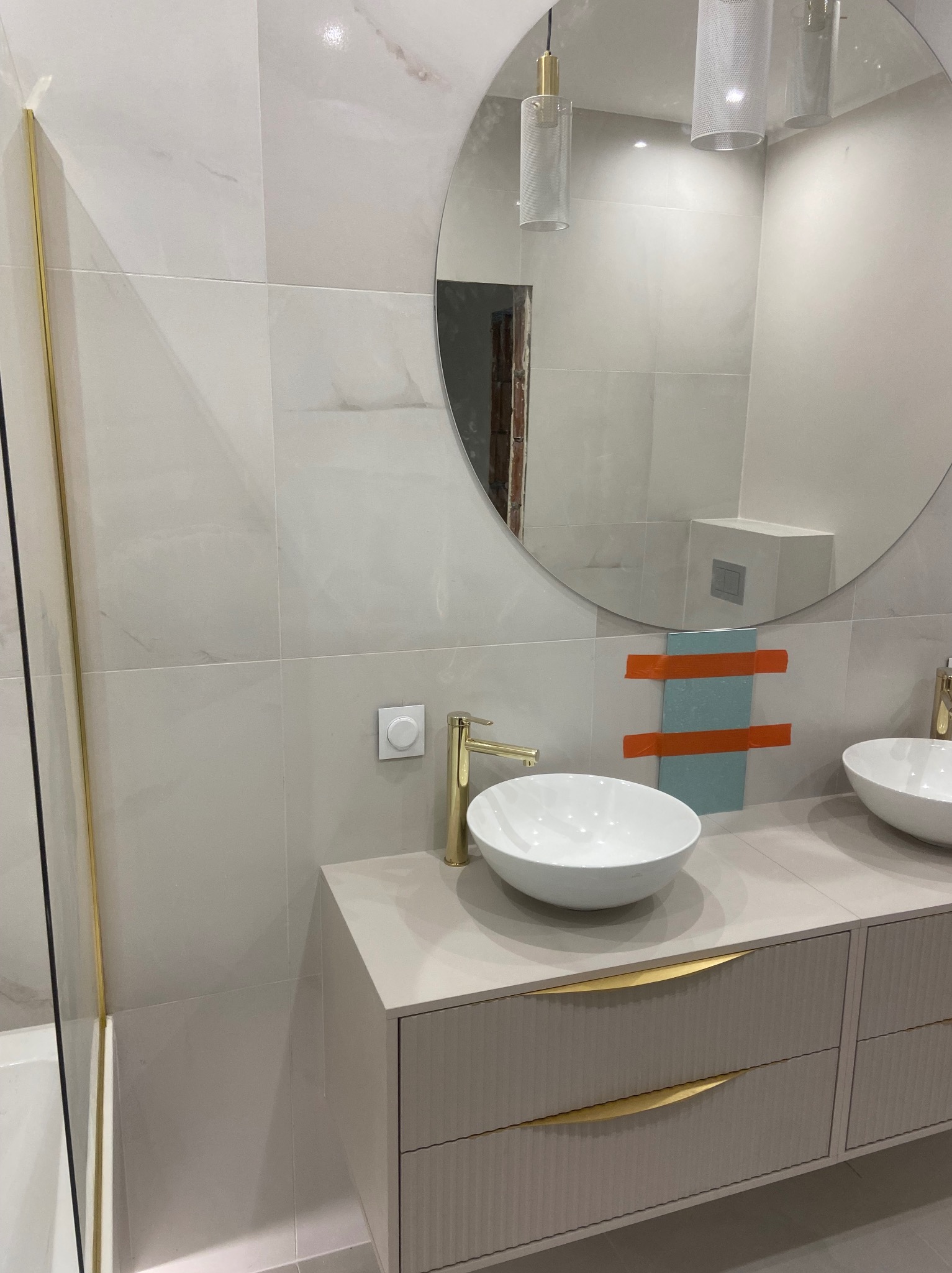 Elegancka łazienka z umywalkami nablatowymi, złotą armaturą i dużym okrągłym lustrem. Widoczne płytki imitujące marmur i szafka z frezowanymi frontami.