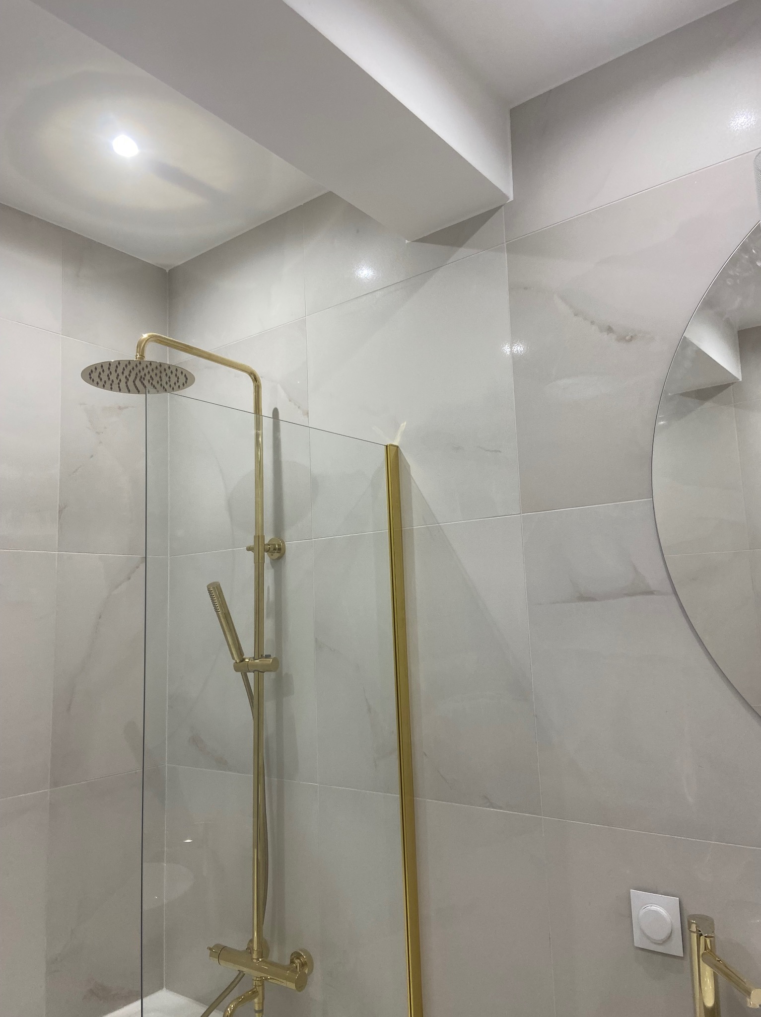 Wykończona łazienka z kabiną prysznicową typu walk-in, złotą armaturą i okrągłym lustrem. Ściany wyłożone płytkami imitującymi marmur.