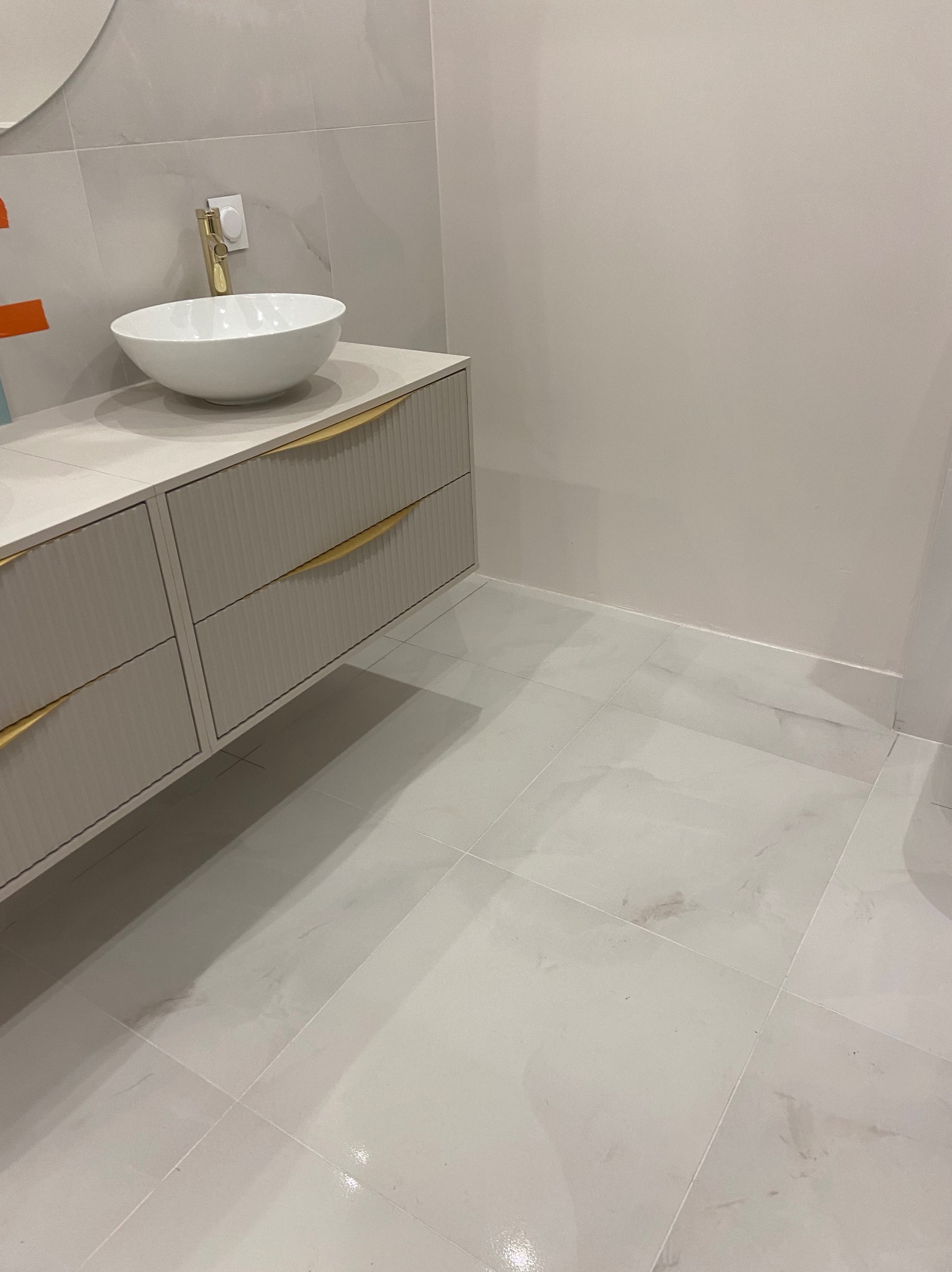 Biała umywalka nablatowa z złotą armaturą na szafce z ryflowanymi frontami i złotymi uchwytami. Jasne płytki podłogowe i ścienne imitujące marmur.