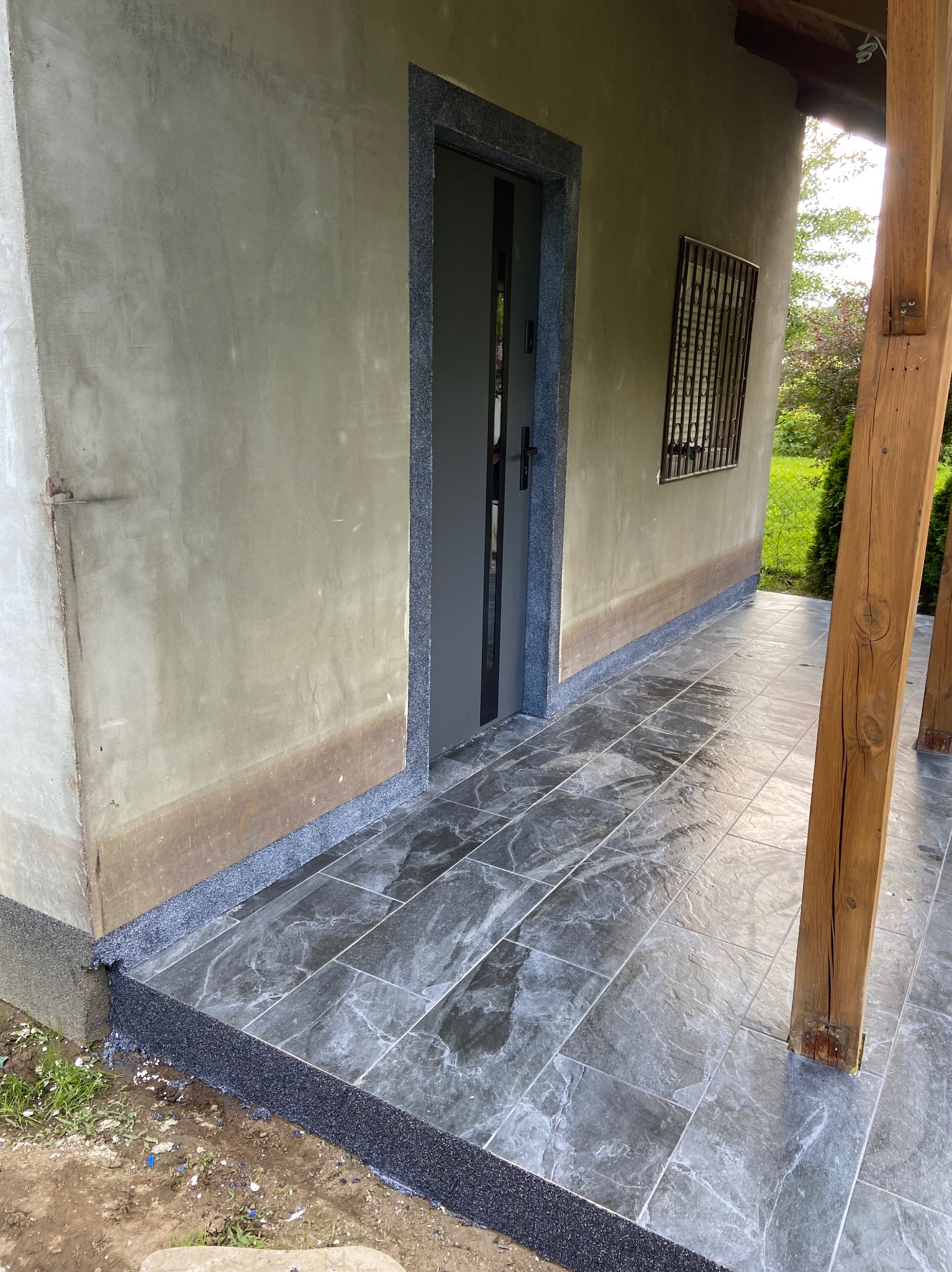 Wykończony taras z szarymi płytkami imitującymi marmur, obramowany czarnym, drobnym kamieniem. Szara stolarka drzwiowa i drewniane słupy podtrzymujące zadaszenie.