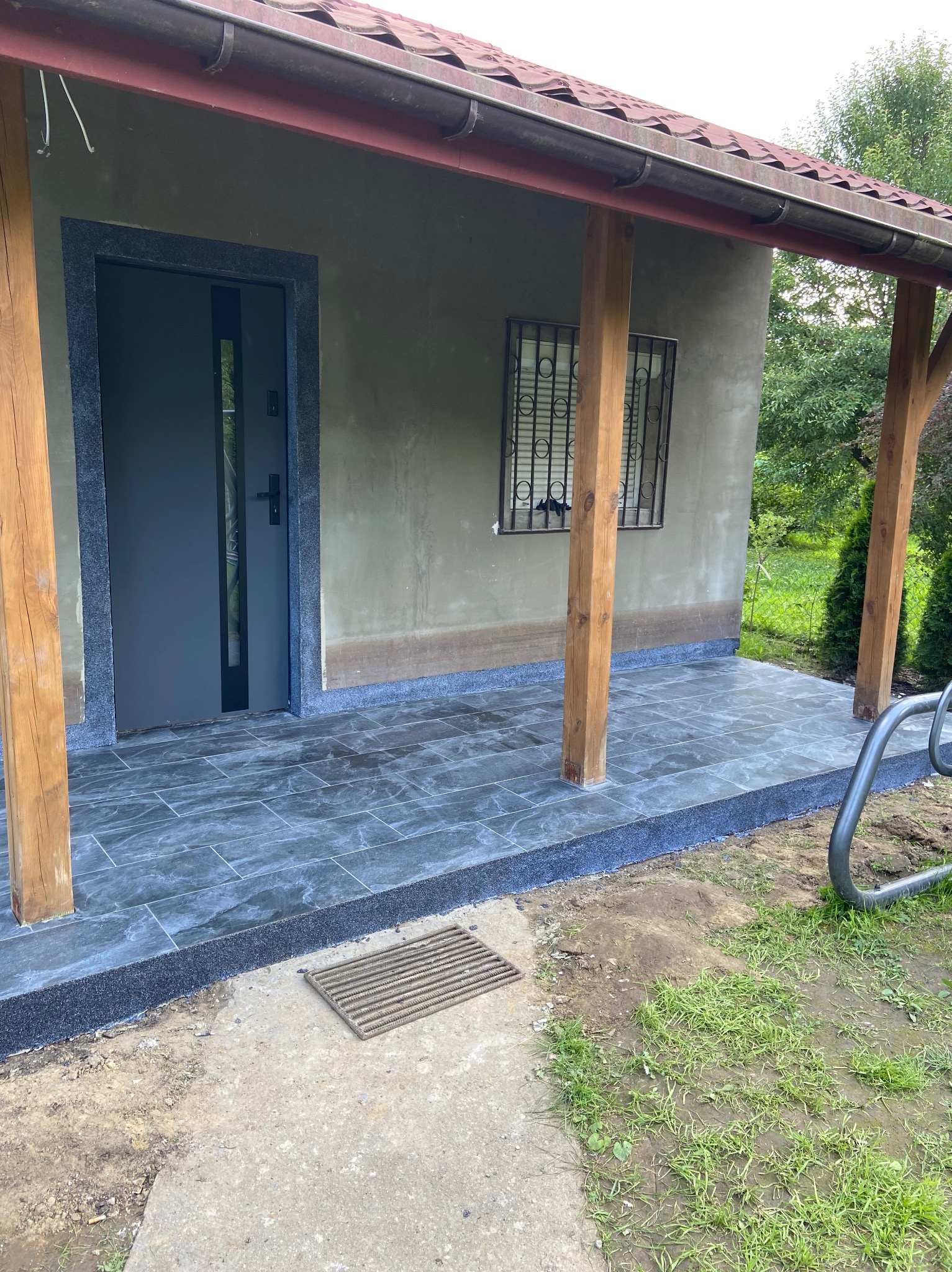 Zadaszone wejście do budynku z nową, ciemną, marmurkową posadzką z płytek. Widoczne drewniane podpory, okno z kratą i szare drzwi wejściowe.