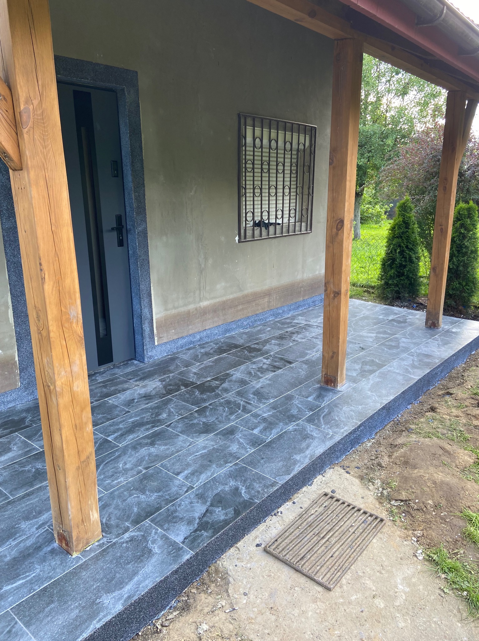 Zadaszony taras z szarych płytek imitujących kamień, obramowany ciemnym gresem. Widoczne drewniane słupy podtrzymujące dach i metalowa krata w oknie.