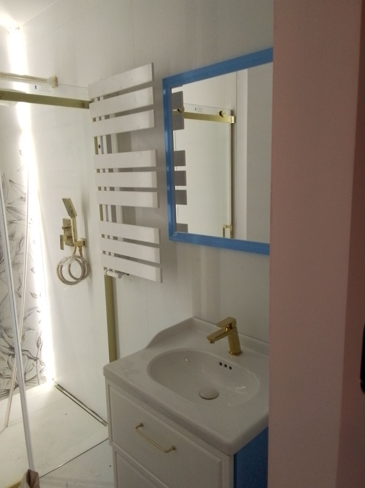 Nowoczesna łazienka z białą szafką umywalkową, złotą armaturą i grzejnikiem drabinkowym. Lustro w niebieskiej ramie odbija kabinę prysznicową. Minimalistyczny design.