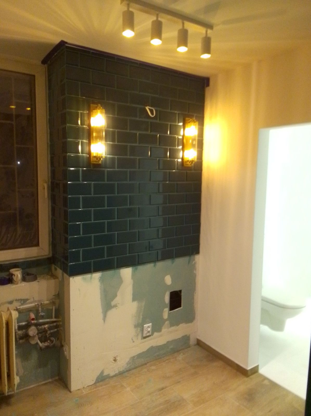 Ściana w łazience wyłożona ciemnymi, błyszczącymi płytkami, z zamontowanymi kinkietami. Dolna część ściany przygotowana do dalszych prac wykończeniowych.