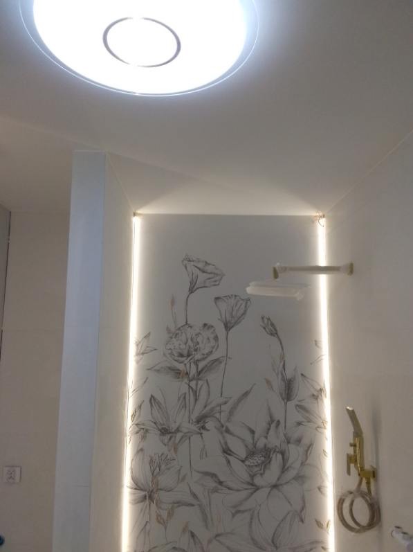 Biała łazienka z podświetlaną ścianą w kwiatowe wzory, złota armatura i okrągła lampa sufitowa. Nowoczesny design z akcentem natury i delikatnym oświetleniem.