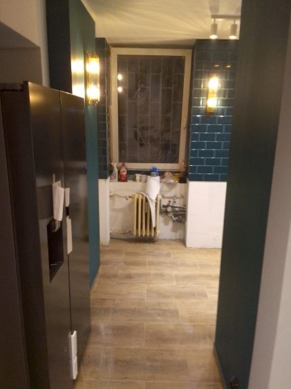 Remont kuchni w trakcie: położona podłoga z płytek imitujących drewno, ściany z niebieskiej glazury, grzejnik i instalacje przed zabudową. Widok z perspektywy wejścia.