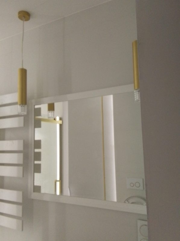 Nowoczesna łazienka z białymi ścianami, lustrem z podświetleniem LED i złotymi lampami wiszącymi. Minimalistyczny design z grzejnikiem drabinkowym.