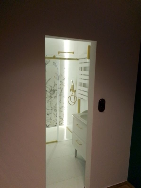 Nowoczesna łazienka z kabiną prysznicową ze złotymi detalami, białą umywalką i grzejnikiem drabinkowym, widziana zza ściany. Minimalistyczny design.