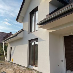 KRZYSZTOF KWAŚNIAK - Nowoczesny dom jednorodzinny z jasną elewacją, brązowymi oknami i dachem, widok z boku na świeżo otynkowaną ścianę i przygotowany teren wokół budynku.