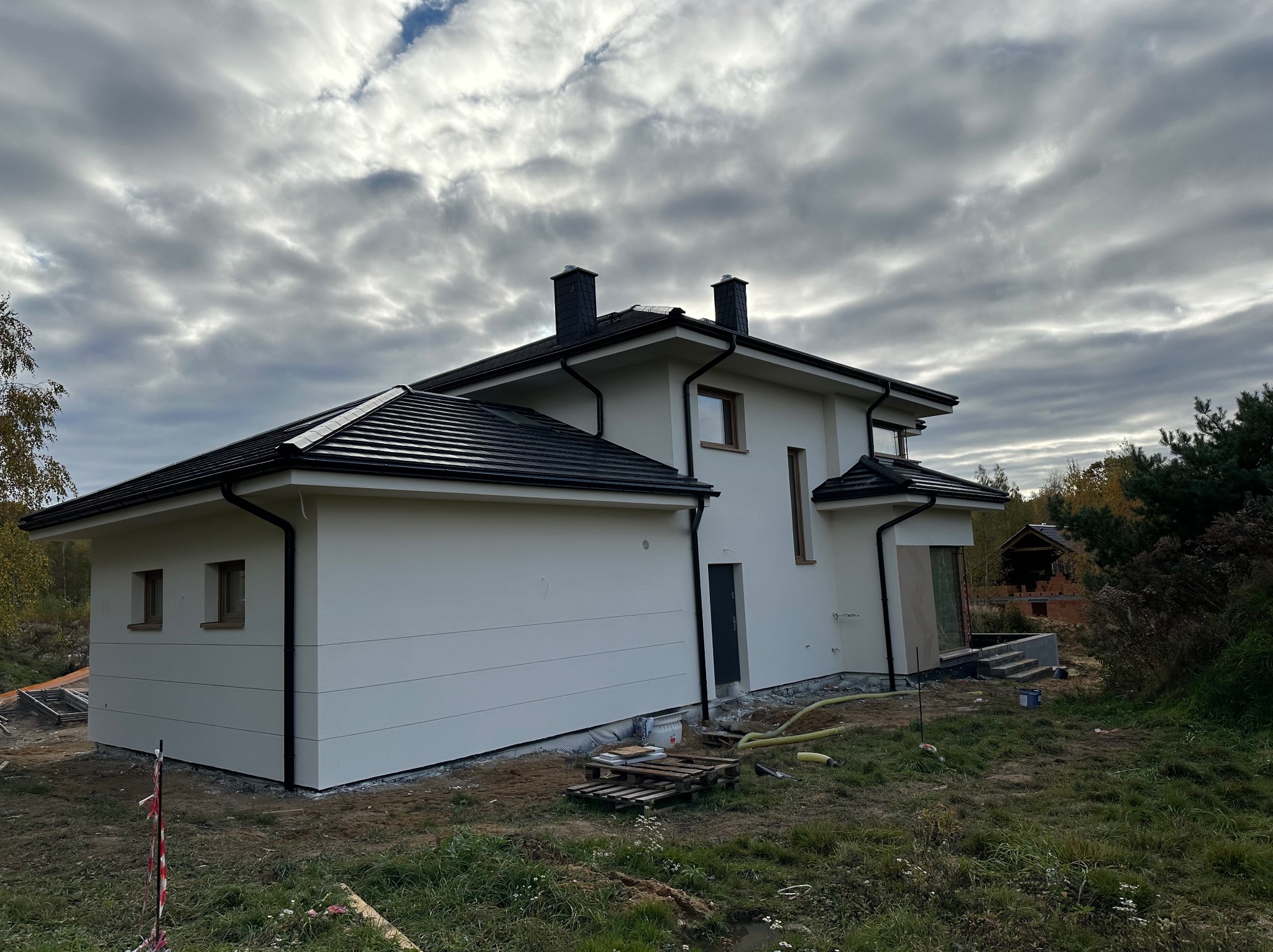 Nowoczesny dom jednorodzinny z jasną, gładką elewacją i ciemną dachówką, widok z boku, teren wokół domu w trakcie prac wykończeniowych, widoczne palety i węże.