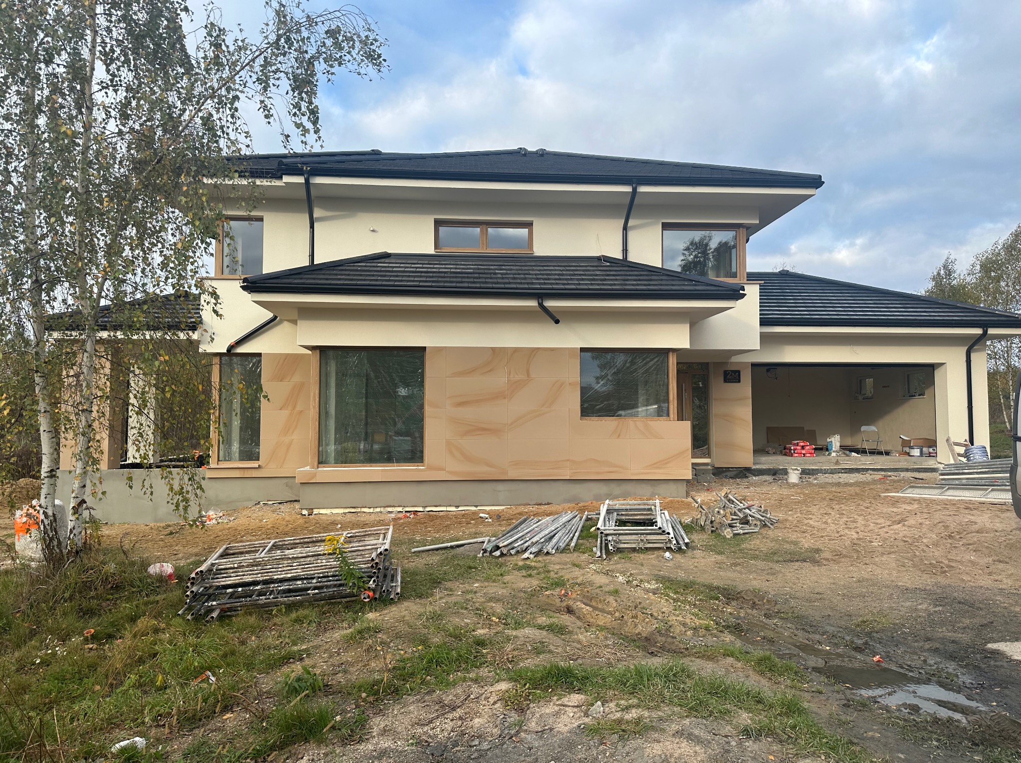 Dwupiętrowy dom w trakcie budowy z beżową elewacją, częściowo wykończoną płytami imitującymi piaskowiec, czarnym dachem i rozłożonymi rusztowaniami na nierównym terenie.