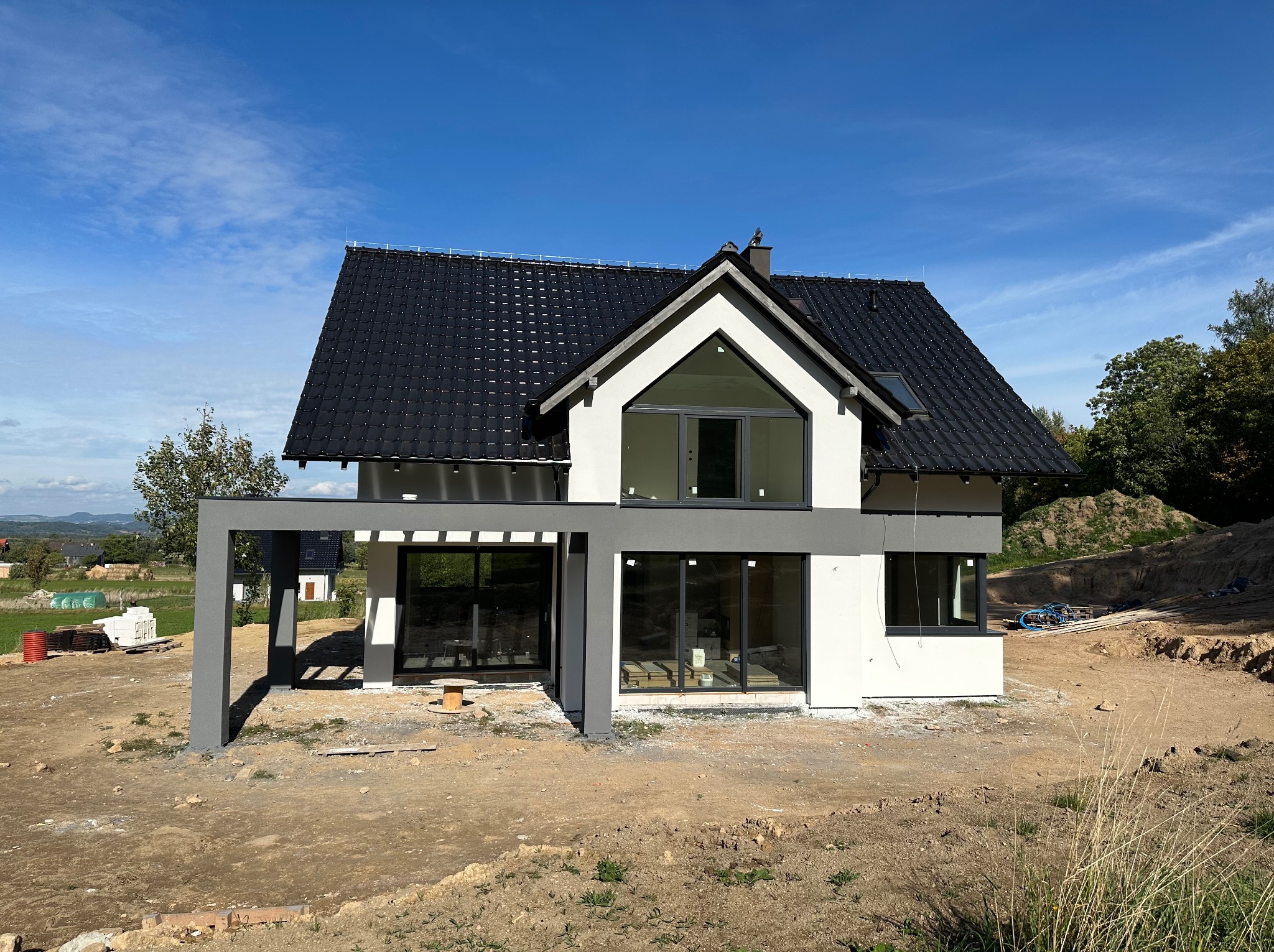 Nowoczesny dom jednorodzinny z płaskim dachem i szarą pergolą, elewacja w kolorze białym i szarym, widoczne okna z czarnymi ramami, teren wokół domu w trakcie prac ziemnych.