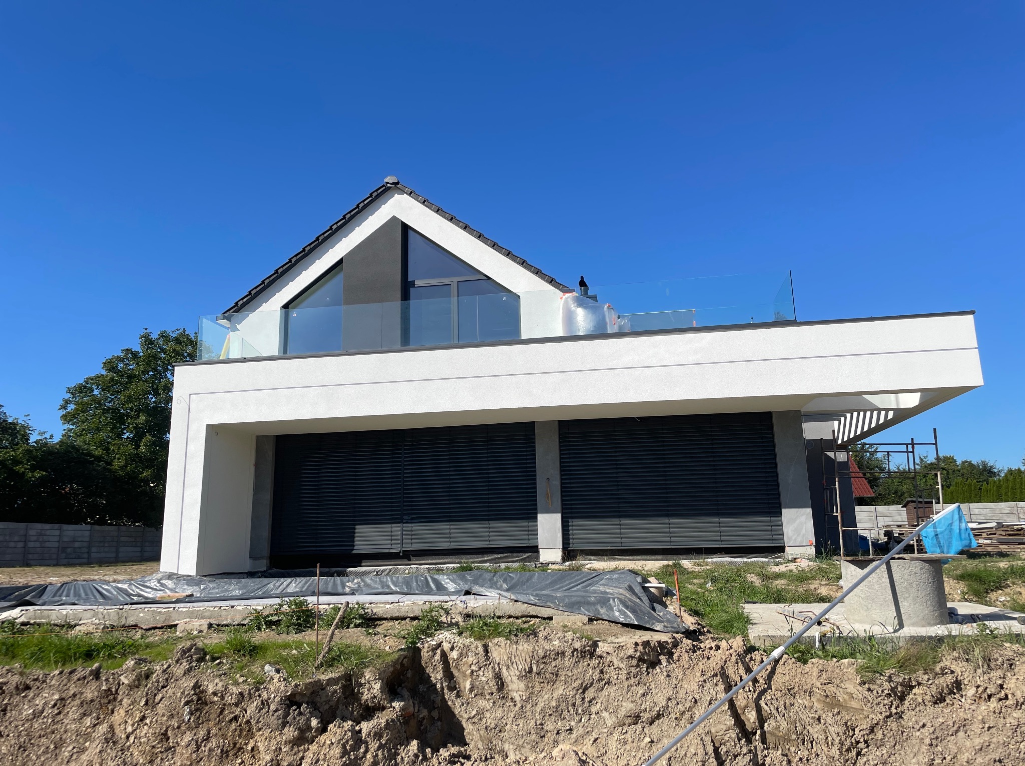 Nowoczesny, dwukondygnacyjny dom z płaskim dachem i szklaną balustradą na górnym tarasie, z widocznymi roletami zewnętrznymi, w trakcie prac wykończeniowych wokół budynku, na tle błękitnego nieba.