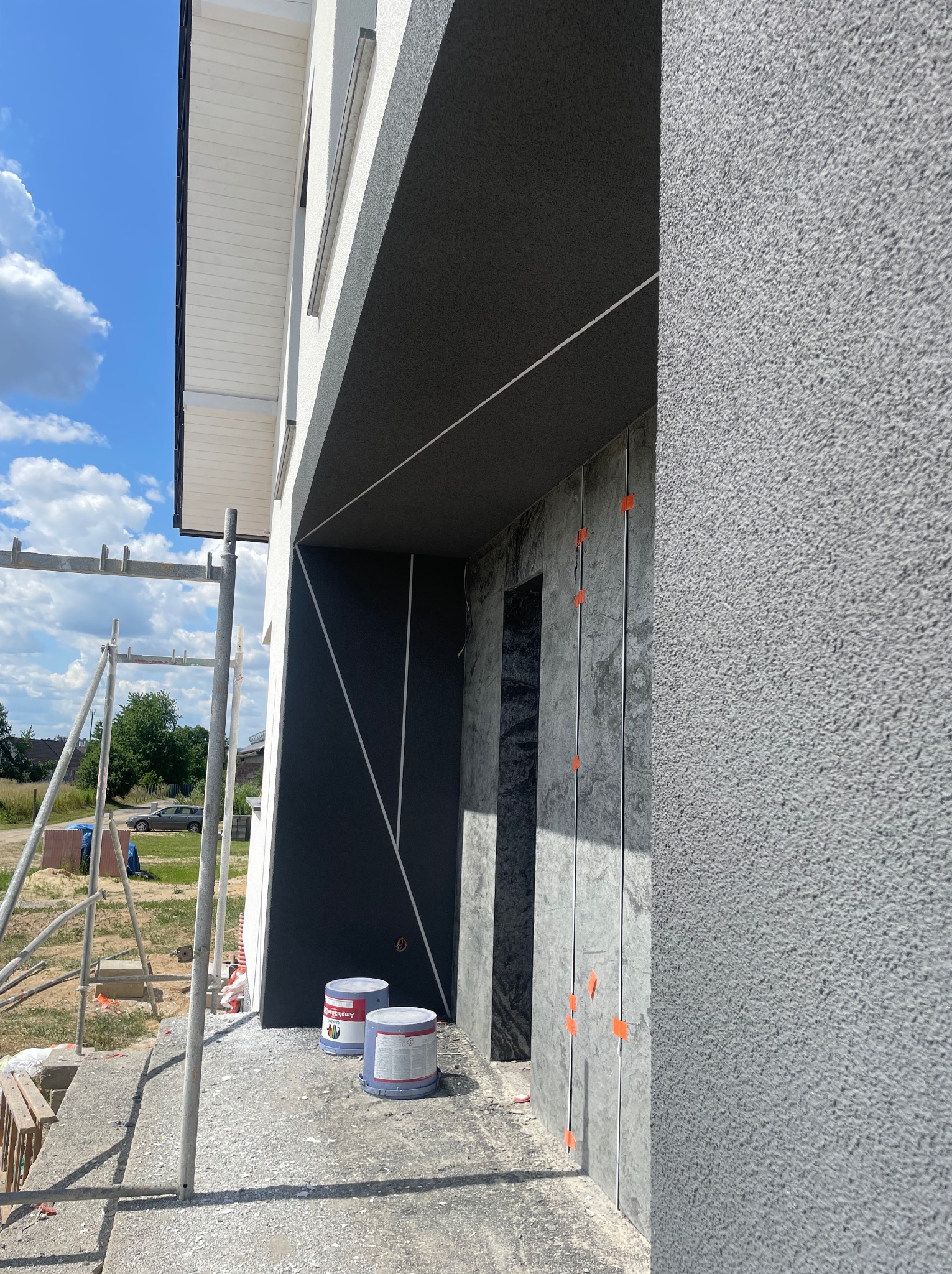 Elewacja domu w trakcie wykończenia: szara struktura tynku, czarne elementy dekoracyjne, widoczne rusztowanie i puszki z farbą AmphiSilan na podłożu.