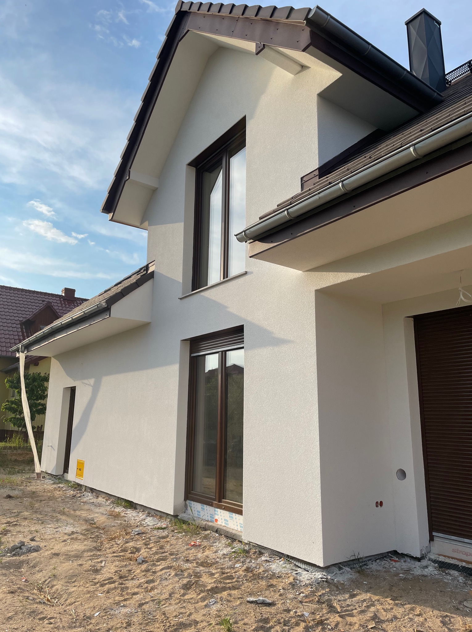 Nowoczesny dom jednorodzinny z jasną elewacją, brązowymi oknami i dachem, widok z boku na świeżo otynkowaną ścianę i przygotowany teren wokół budynku.