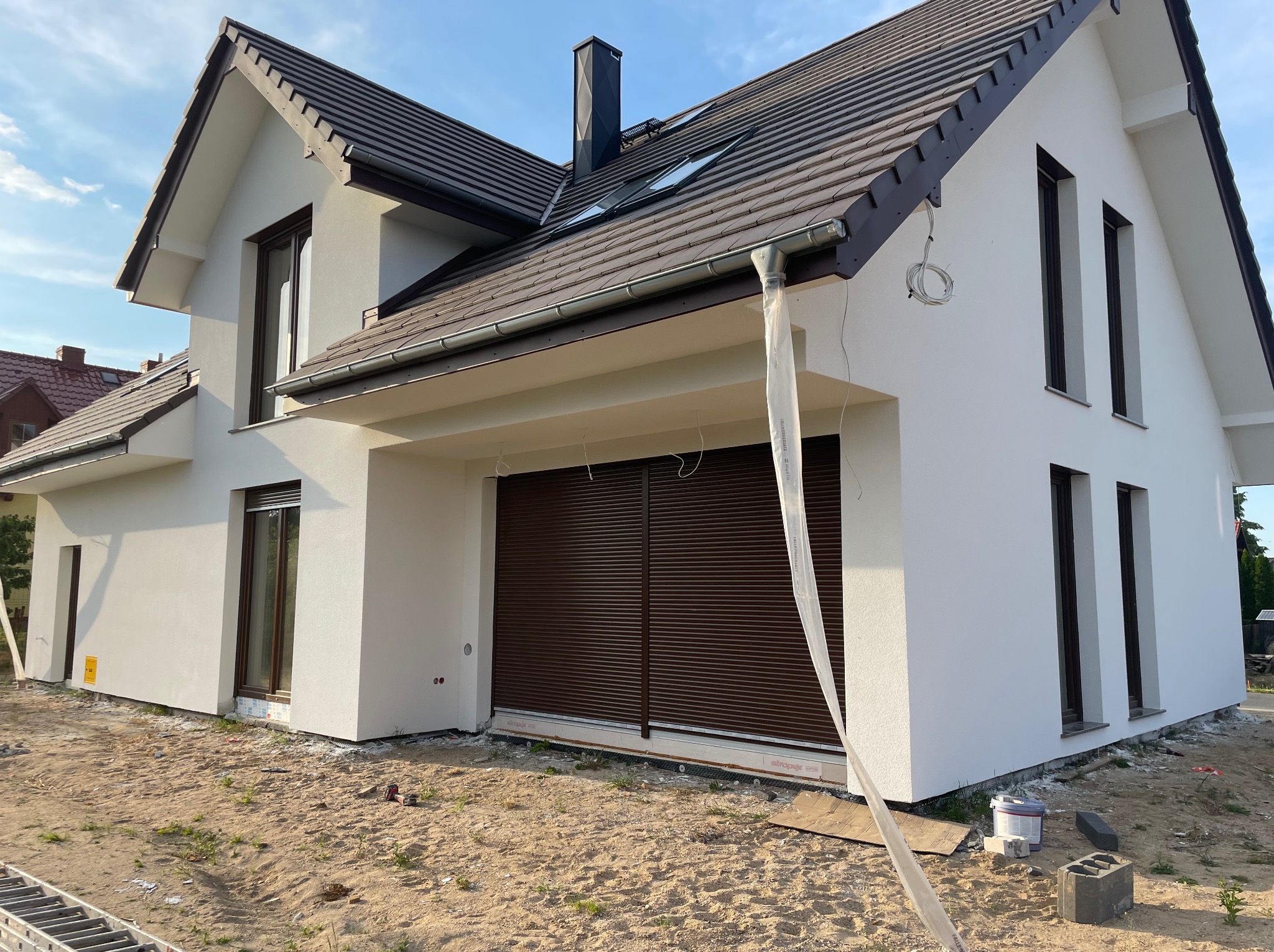 Nowoczesny dom jednorodzinny z jasną elewacją i brązową stolarką okienną, widoczne przygotowania do prac wykończeniowych wokół budynku, w tle błękitne niebo.