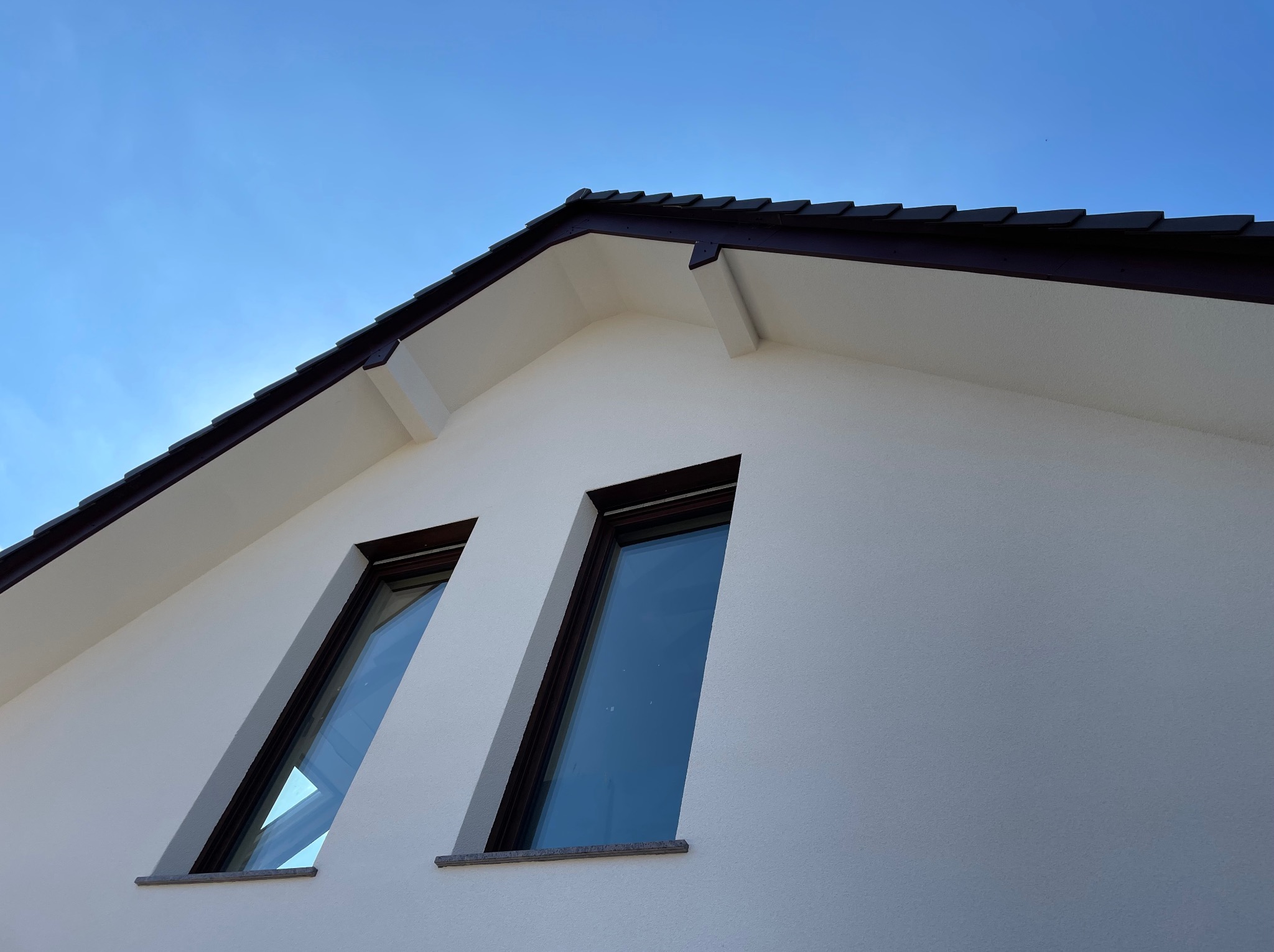 Fragment nowo wykończonej elewacji domu z dwoma oknami na poddaszu, widok z dołu na tle błękitnego nieba.