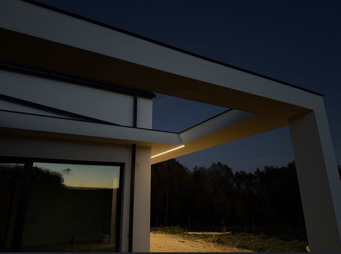 Nowoczesna elewacja domu z oświetleniem LED pod zadaszeniem, widok z dołu na tle wieczornego nieba. Minimalistyczny design, białe ściany i ciemne obramowania okien.