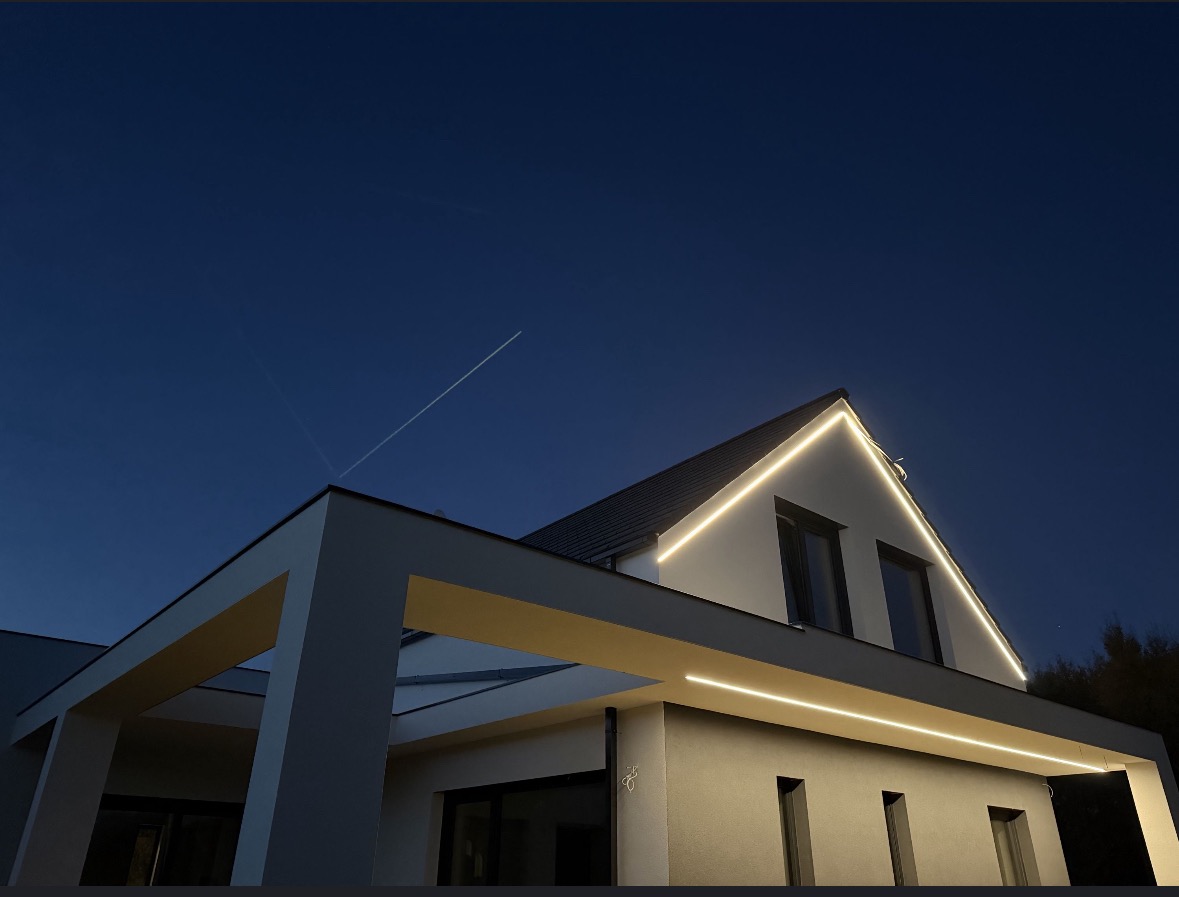 Nowoczesny dom z podświetloną elewacją w odcieniach szarości i bieli, oświetlenie LED podkreśla architekturę budynku na tle ciemniejącego nieba.