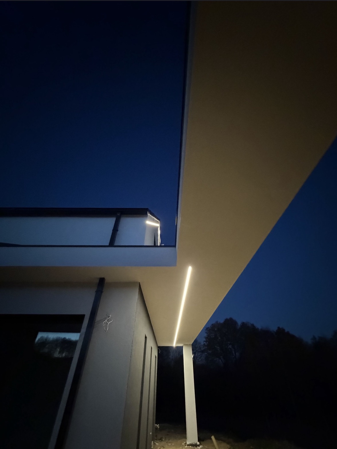 Nowoczesny dom z oświetleniem elewacyjnym LED, nocne ujęcie z dołu. Minimalistyczna architektura, ciemne tło nieba i drzew. Eleganckie wykończenie.