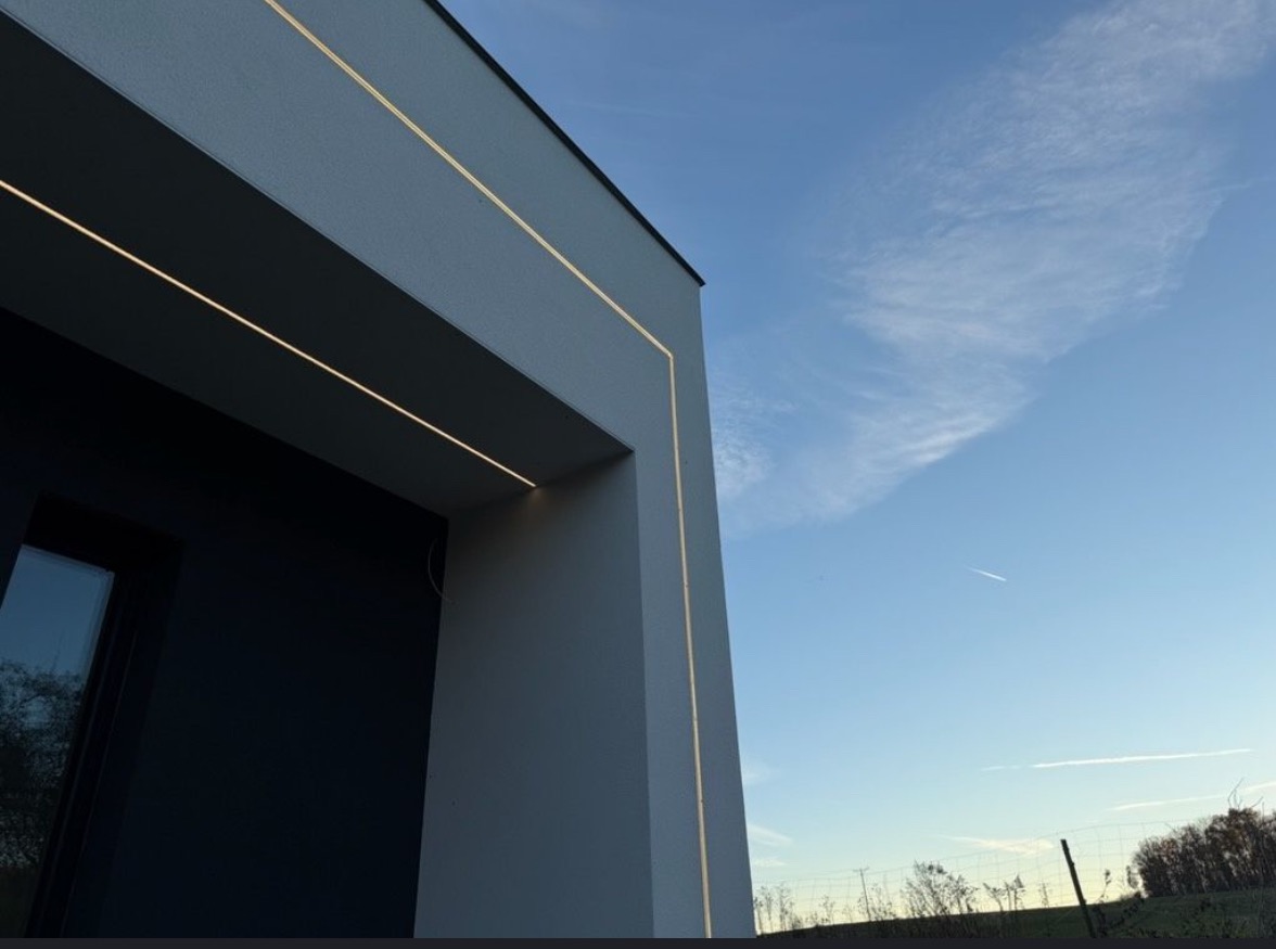 Nowoczesny budynek z szarą elewacją i subtelnym, liniowym oświetleniem LED podkreślającym krawędzie. Błękitne niebo w tle. Ujęcie z dołu.