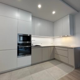 MUBLI - Manufaktura mebli na wymiar - Nowoczesna kuchnia w odcieniach szarości z zabudową na wymiar, obejmującą piekarnik i mikrofalówkę, oświetlenie LED podszafkowe i płytki podłogowe w geometryczny wzór.