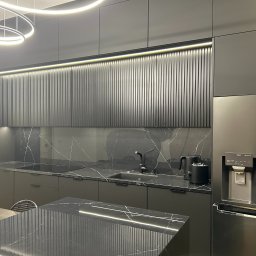 MUBLI - Manufaktura mebli na wymiar - Nowoczesna kuchnia w odcieniach szarości z czarnymi blatami marmurkowymi, podświetlanymi szafkami górnymi z pionowymi żłobieniami oraz wyspą z dodatkowym oświetleniem LED.