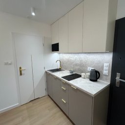 MUBLI - Manufaktura mebli na wymiar - Nowoczesna, jasna kuchnia z szarymi szafkami dolnymi i beżowymi górnymi, złotą armaturą, blatem imitującym marmur, czajnikiem elektrycznym i płytą indukcyjną, oświetlona punktowymi lampami.