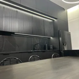 Nowoczesna, czarna kuchnia z marmurowym blatem, oświetleniem LED pod szafkami i designerską lampą sufitową, widziana zza drewnianego stołu z krzesłami o plecionych oparciach.