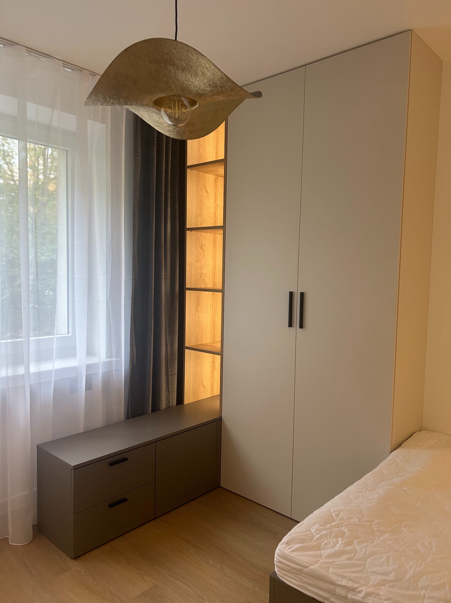 Jasna szafa wnękowa z ciemnymi uchwytami, zintegrowana z otwartymi półkami z podświetleniem, obok okna z białymi i szarymi zasłonami, oraz szafka z szufladami i fragment łóżka.