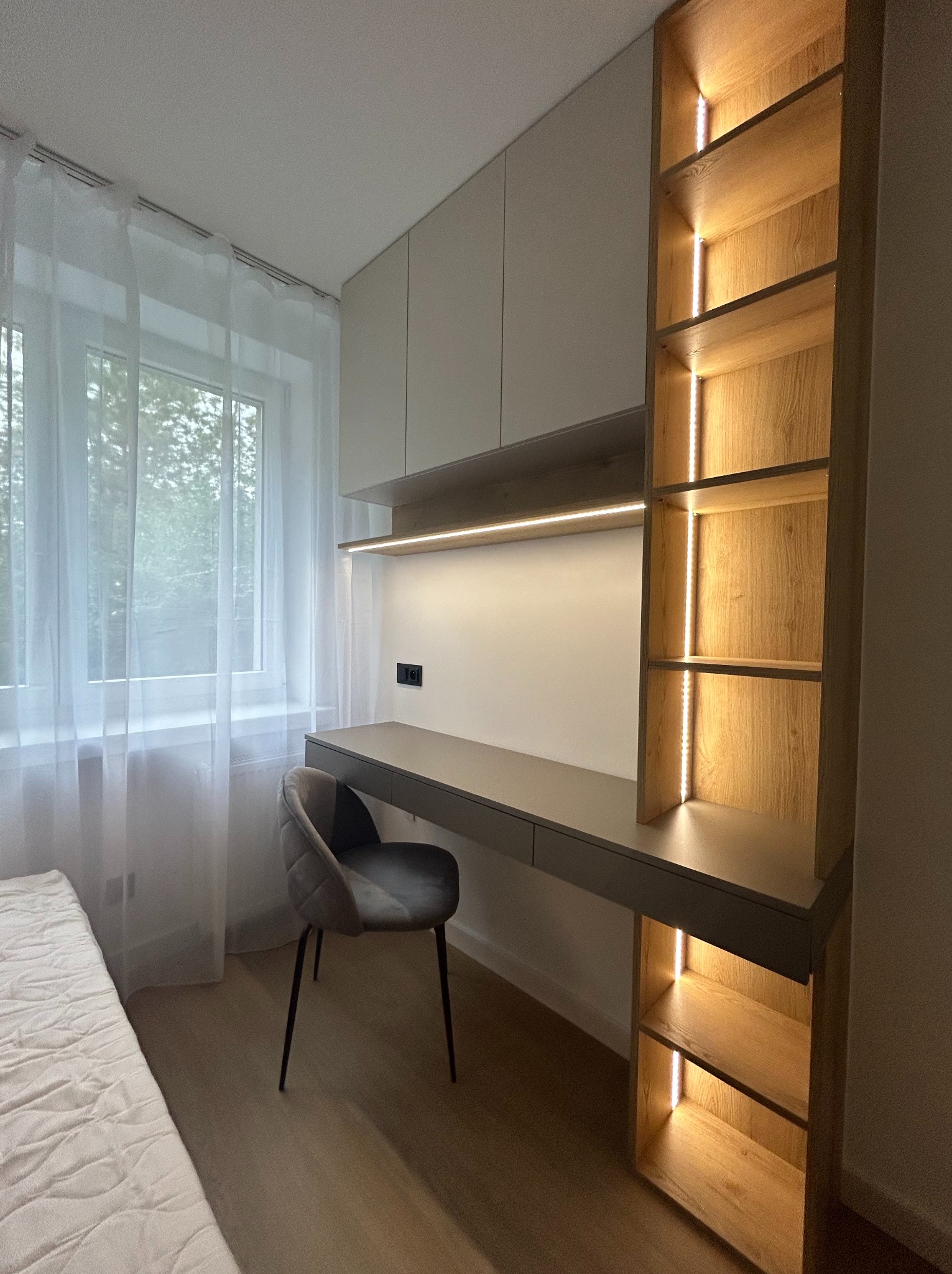 Nowoczesne biurko zawieszone na ścianie z szarym blatem, szarą tapicerowaną krzesłem, podświetlanymi półkami z jasnego drewna i szafkami wiszącymi w jasnych kolorach, w pokoju z zasłoniętym oknem.