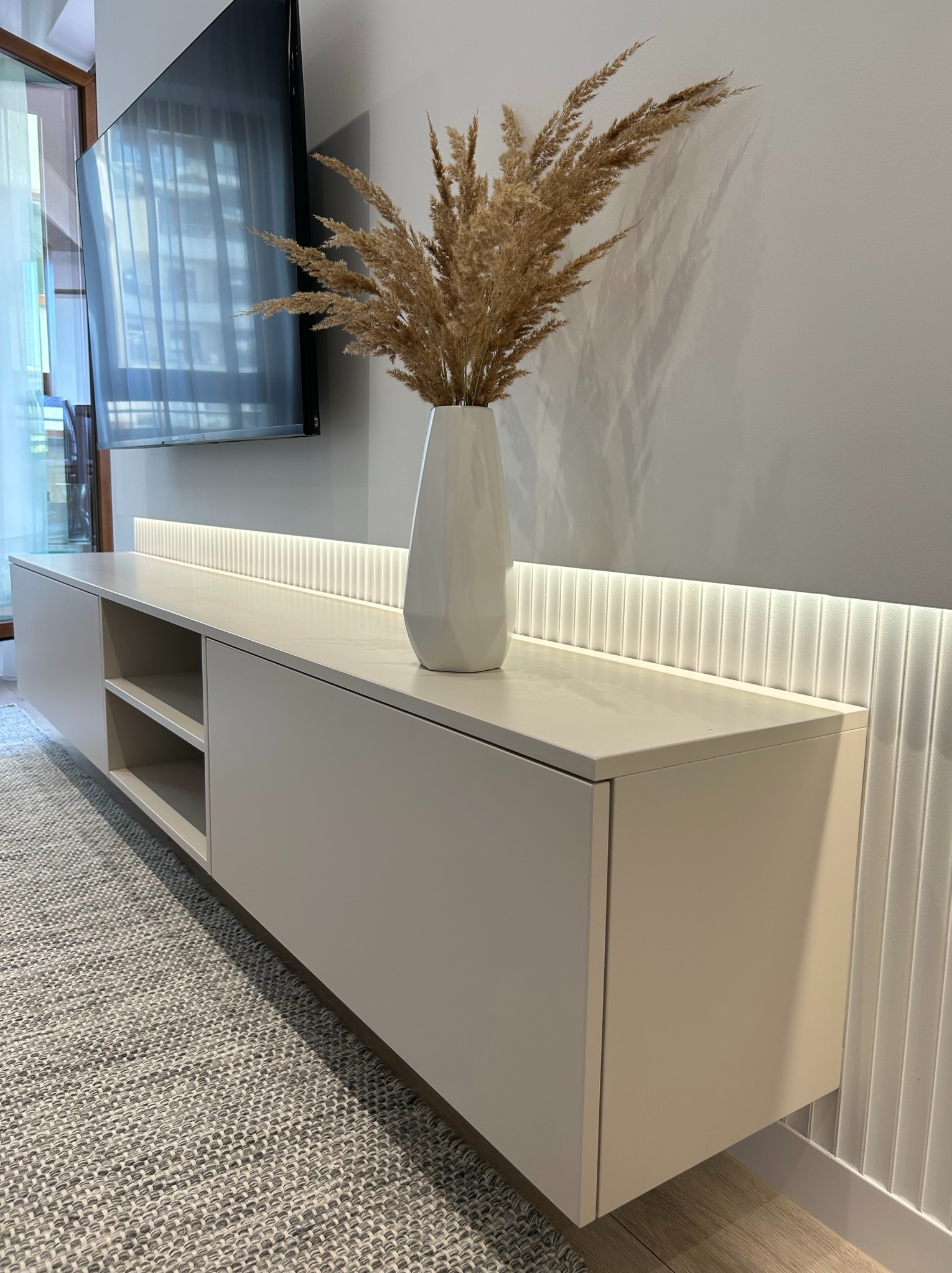 Nowoczesna szafka RTV w odcieniach beżu, podwieszana, z otwartymi półkami i zamkniętymi szafkami, oświetlona listwą LED, z dekoracyjnym wazonem z suszonymi trawami, na tle ściany z pionowymi...