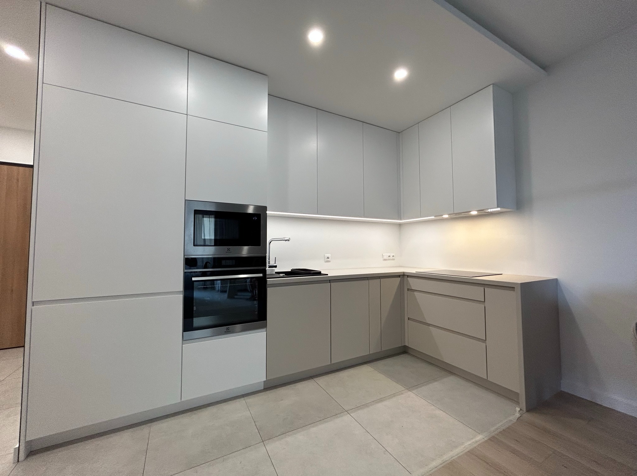 Nowoczesna kuchnia w odcieniach szarości z zabudową na wymiar, obejmującą piekarnik i mikrofalówkę, oświetlenie LED podszafkowe i płytki podłogowe w geometryczny wzór.